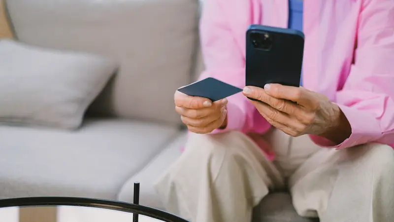 Personne âgée assise sur un canapé, un smartphone dans une main et une carte bancaire dans l'autre