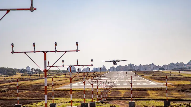 Avion à hélices au-dessus d'une piste bordée de rampes de balisage rouges et blanches, une ville en arrière-plan