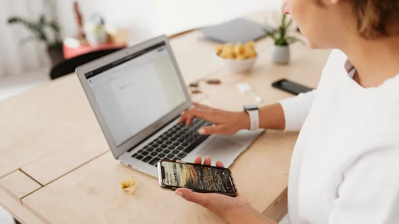 Femme assise à une table en bois, une main sur le clavier d'un ordinateur portable, un smartphone dans l'autre main