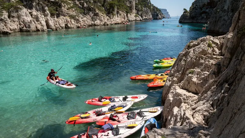 Kayaks colorés amarrés contre la falaise d'une calanque aux eaux turquoise, baigneurs et pagayeurs au loin