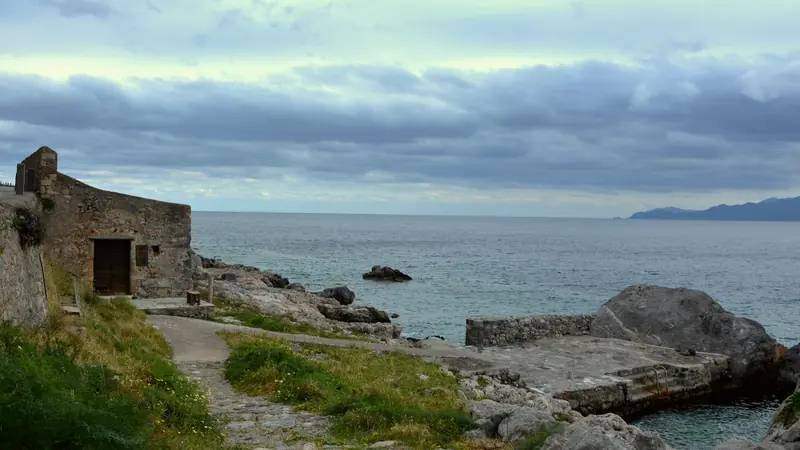 Petit bâtiment de pierre au bord d'un chemin côtier rocheux, face à la mer sous un ciel nuageux