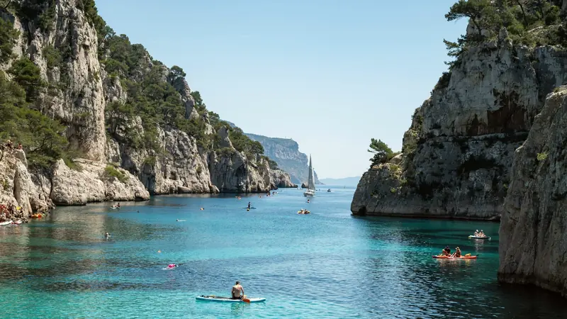Calanque aux falaises calcaires boisées de pins, eau turquoise parcourue de paddles, de kayaks et d'un voilier