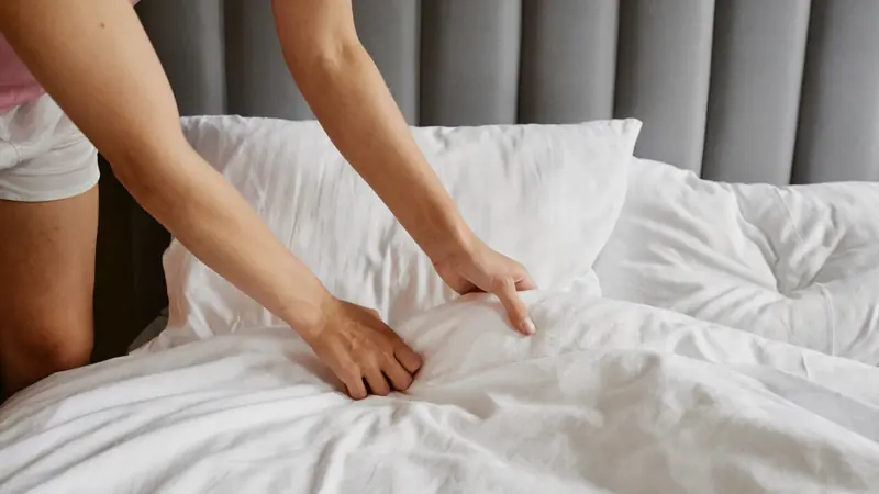 Mains arrangeant un oreiller et une couette blanche sur un lit fraîchement refait