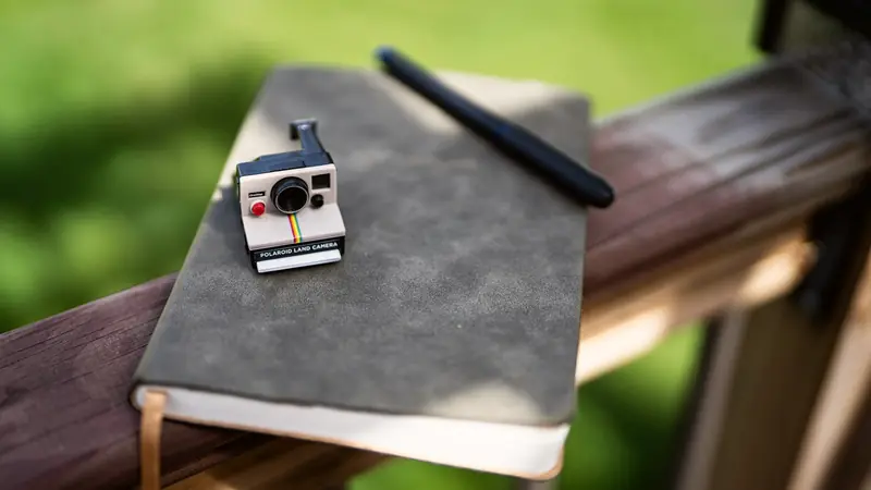 Carnet à couverture grise, stylo noir et petit appareil photo instantané posés sur une rambarde en bois