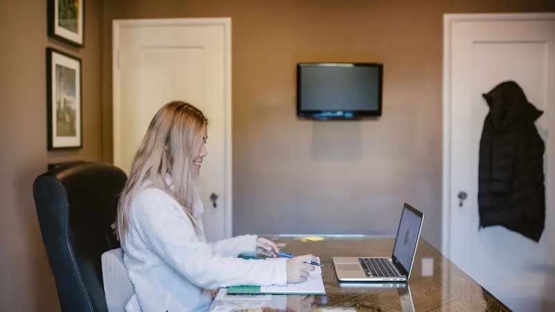 Jeune femme assise à une grande table, prenant des notes sur un cahier devant un ordinateur portable ouvert