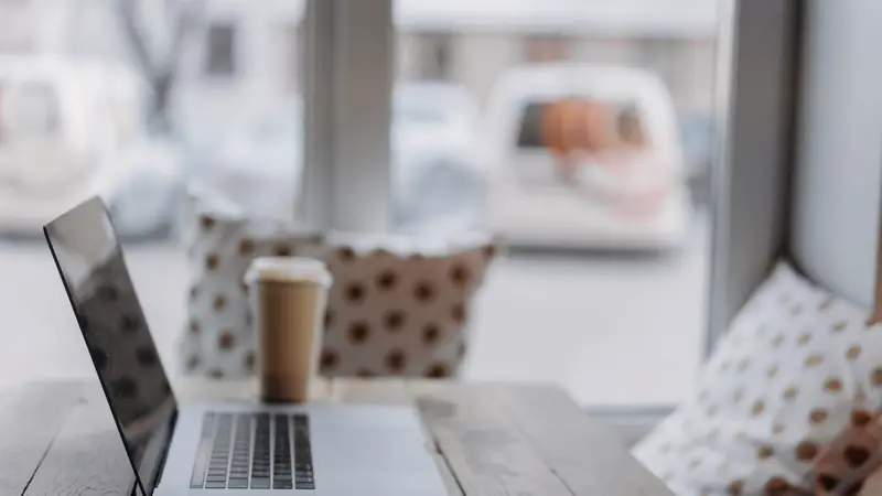Ordinateur portable ouvert sur une table en bois près d'une fenêtre, avec un gobelet de café