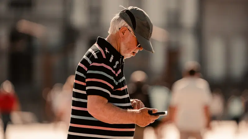 Homme aux cheveux blancs, casquette et polo rayé, consultant son smartphone dans une rue ensoleillée