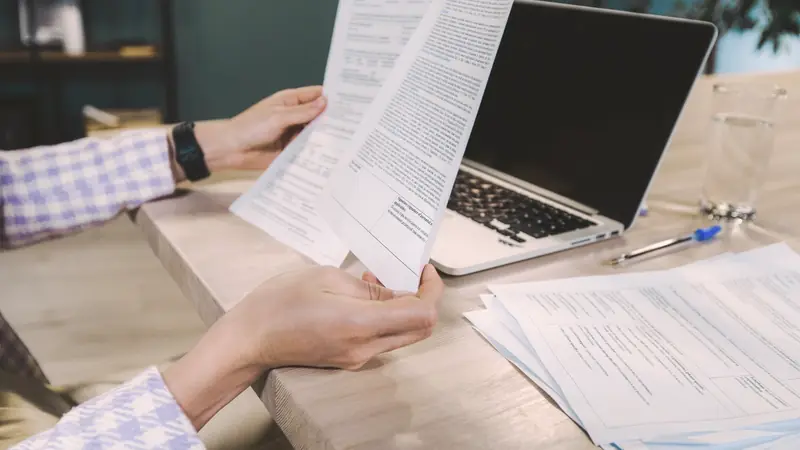 Mains tenant des feuilles imprimées devant un ordinateur portable ouvert, sur une table en bois