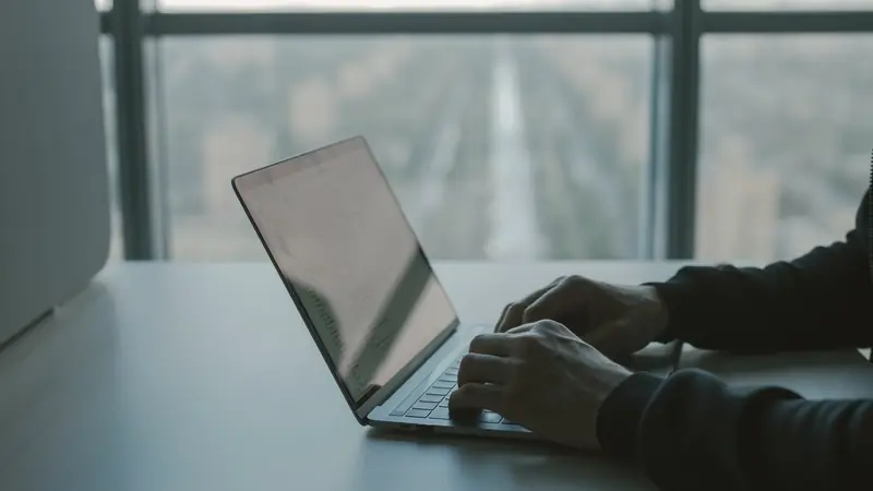 Mains tapant sur un ordinateur portable posé sur un bureau, devant une baie vitrée donnant sur une ville